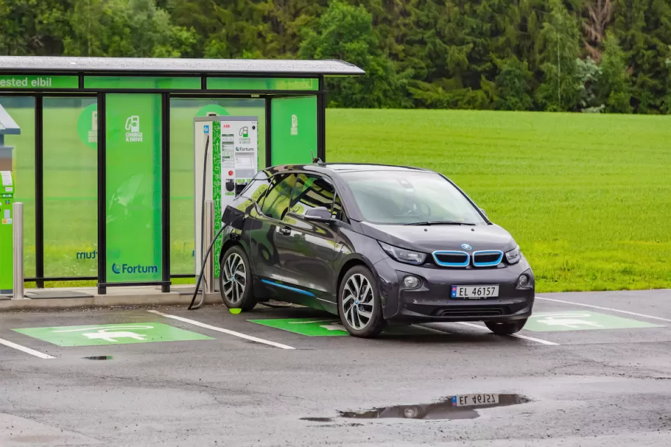 Small grey sedan charging at outdoor electric car charging station with green kiosk and green forest in background in Norway.